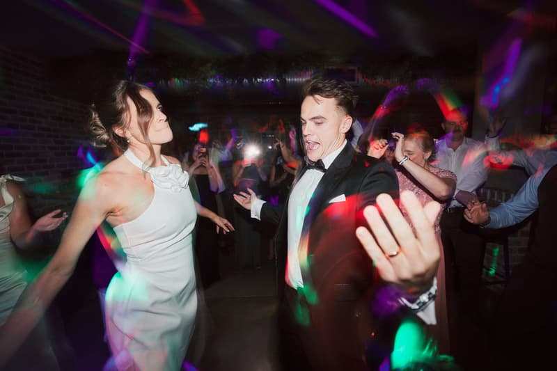Bride Ashleigh in a white dress and groom James in a tuxedo dance energetically at the reception with guests visible in the background at Sandstone Point Hotel — The Cellar.