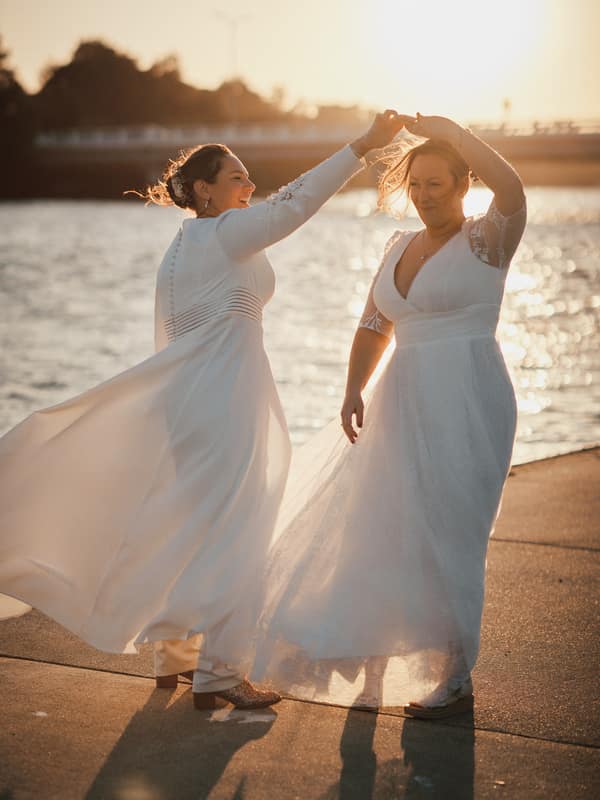 Ashleigh and Lorin, both brides in wedding dresses, dance together near the water at Sandstone Point Hotel during their couple portraits session at sunset.