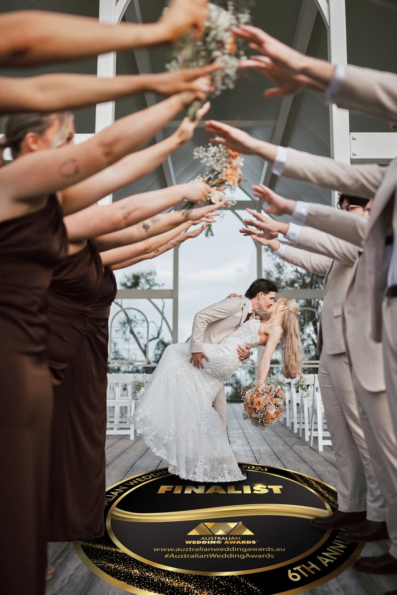Morgan and Joel's dip kiss under a bridal party arch at the Sandstone Point Hotel chapel, with the 2026 Australian Wedding Awards Finalist roundel as an aisle decal