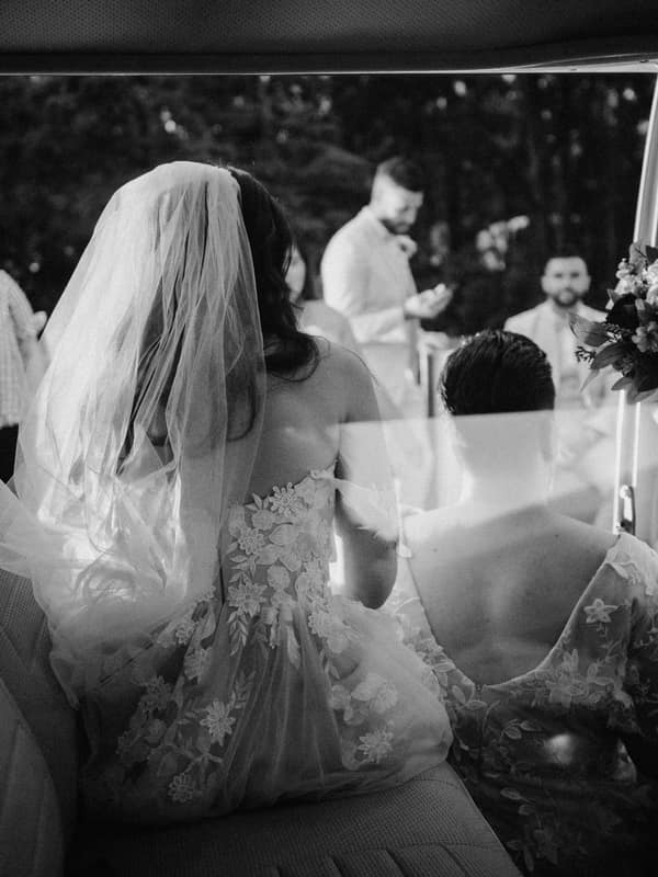 The bride and another woman in floral dresses sit inside a vehicle with the door open, facing a group of people standing outside in a wooded outdoor setting.