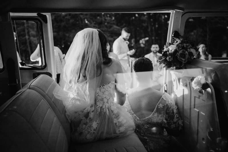 The bride and another woman in floral dresses sit inside a vehicle with the door open, facing a group of people standing outside in a wooded outdoor setting.