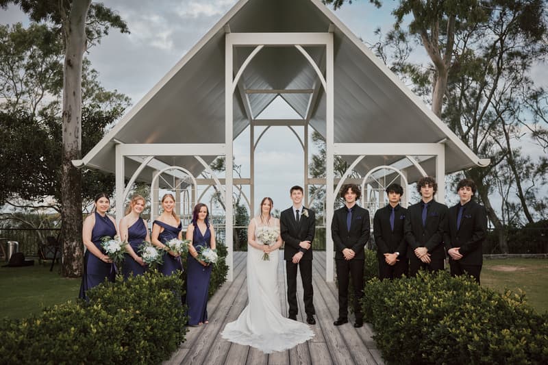 Terion the bride in a white gown and Tyler the groom in a black suit stand together under a white outdoor pavilion at Sandstone Point Hotel, flanked by four bridesmaids in navy dresses holding bouquets on the left and five groomsmen in black suits with navy ties on the right.