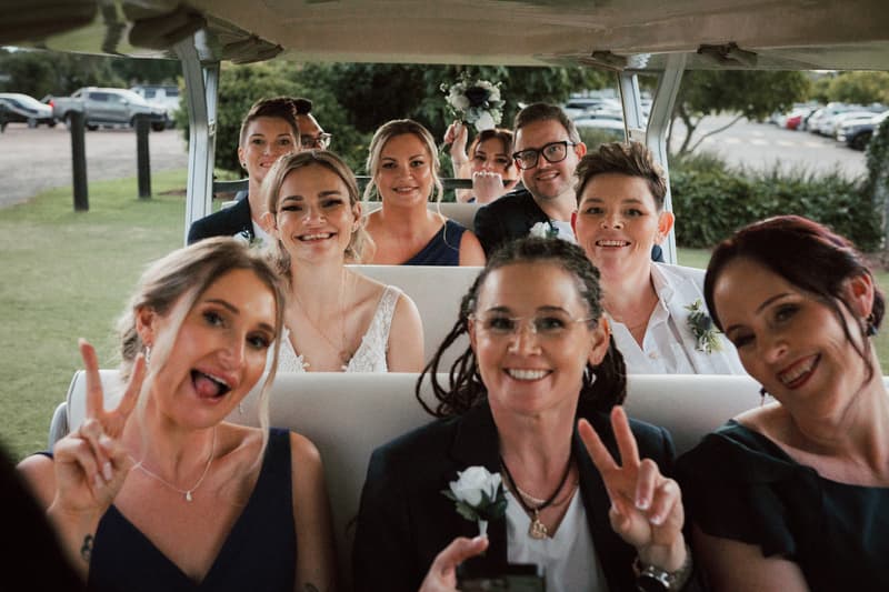 Mandy and Selina with their wedding party seated in a golf cart at Sandstone Point Hotel, some making peace signs and smiling at the camera.