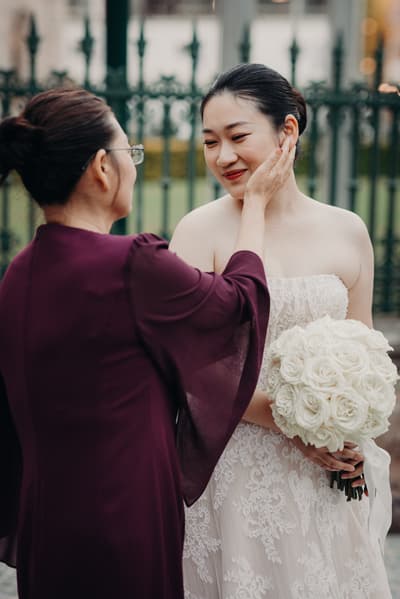 The bride Aria, holding a bouquet of white roses, is gently touched on the cheek by an older woman wearing glasses and a dark purple dress, outside the. Cheap photo booth hire Brisbane by She Said Yes.