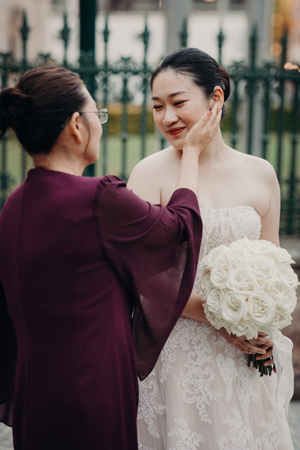 The bride Aria, holding a bouquet of white roses, is gently touched on the cheek by an older woman wearing glasses and a dark purple dress, outside the Queensland Parliament building.