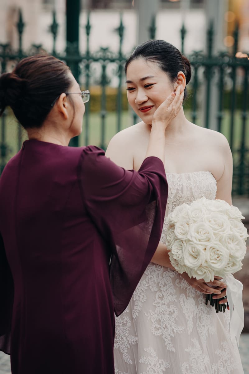 The bride Aria, holding a bouquet of white roses, is gently touched on the cheek by an older woman wearing glasses and a dark purple dress, outside the. Elopement photography Brisbane by She Said Yes.