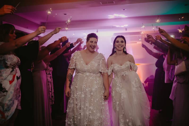 Two brides in lace wedding dresses hold hands and smile while guests on both sides hold sparklers overhead indoors.