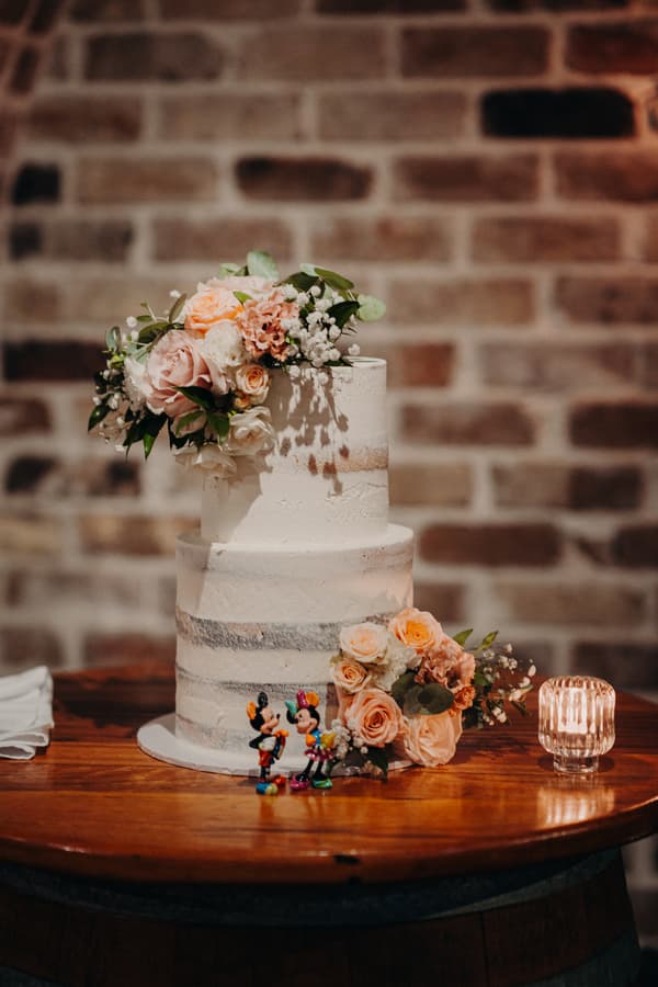 Two-tiered white wedding cake decorated with pastel flowers and two small Mickey and Minnie Mouse figurines on a wooden table at Sandstone Point Hotel.