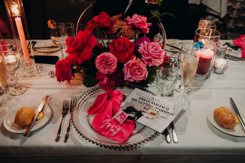Reception table setting with floral centerpiece, candles, champagne glass, bread rolls, and a wedding announcement card for Arran and Jacquelyne. Wedding fair season Brisbane by She Said Yes.