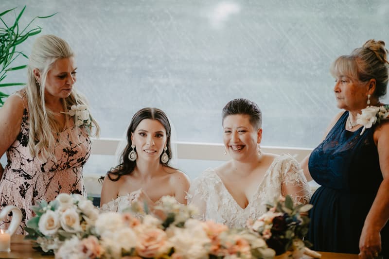 Two brides seated at a table with floral arrangements, flanked by two older women standing on either side, all dressed in formal attire.
