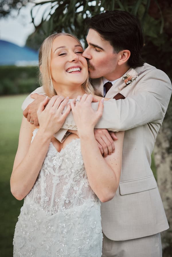 Morgan and Joel embrace outdoors at Sandstone Point Hotel during couple portraits, with Joel kissing Morgan on the cheek.