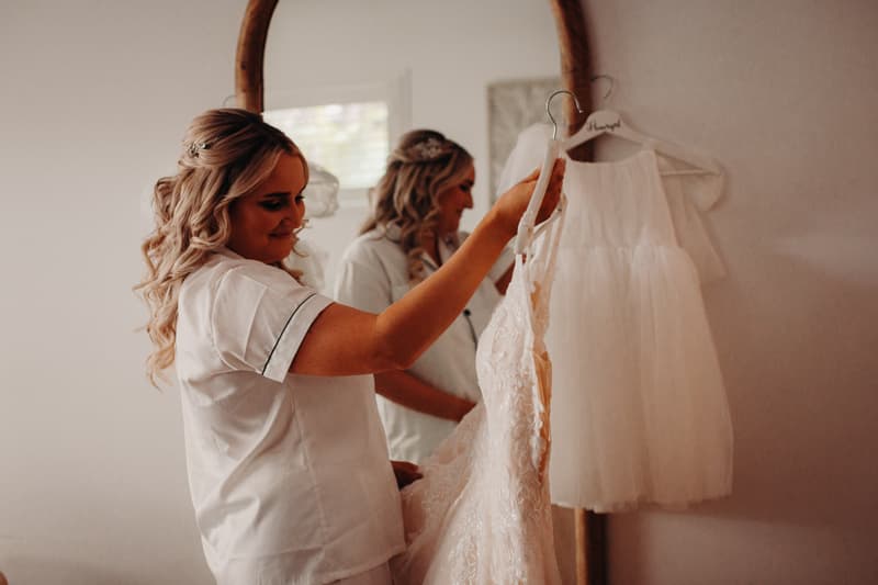 The bride Chloe holds her wedding dress on a hanger while looking at it in front of a mirror during bridal preparation.