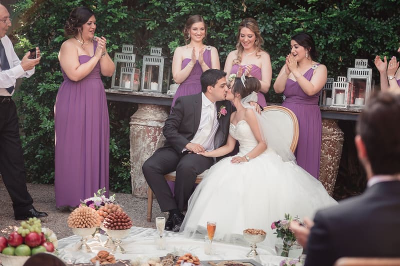 Pasha and Maryam share a kiss seated in front of a table with food and drinks, surrounded by bridesmaids in purple dresses and guests applauding. Full day wedding photography by She Said Yes.