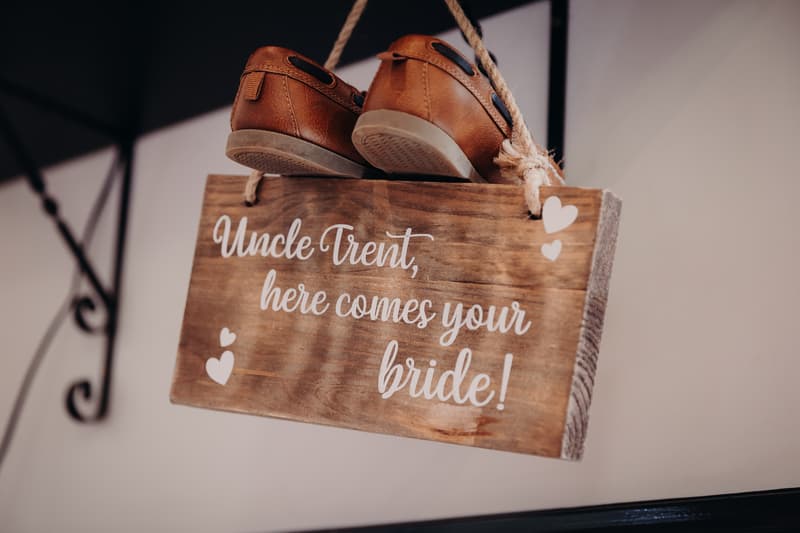 A wooden sign hanging by rope with a pair of brown shoes on top, reading 'Uncle Trent, here comes your bride!'