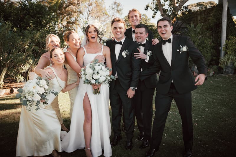 Bride Ashleigh in a white dress holding a bouquet stands with groom James in a black tuxedo and their bridal party of bridesmaids in cream dresses and groomsmen in black tuxedos posing outdoors at Sandstone Point Hotel.