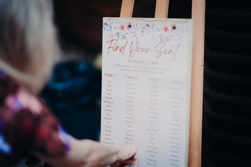 A guest looks at the seating chart displayed on an easel at the reception stage of Yabbaloumba Retreat — The Shed, showing table assignments including the Bridal Party Table and numbered tables.