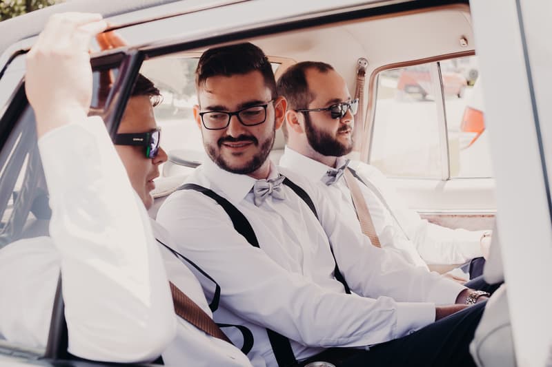 Three groomsmen dressed in white shirts, bow ties, and suspenders sit inside a car at Ocean View Estates.