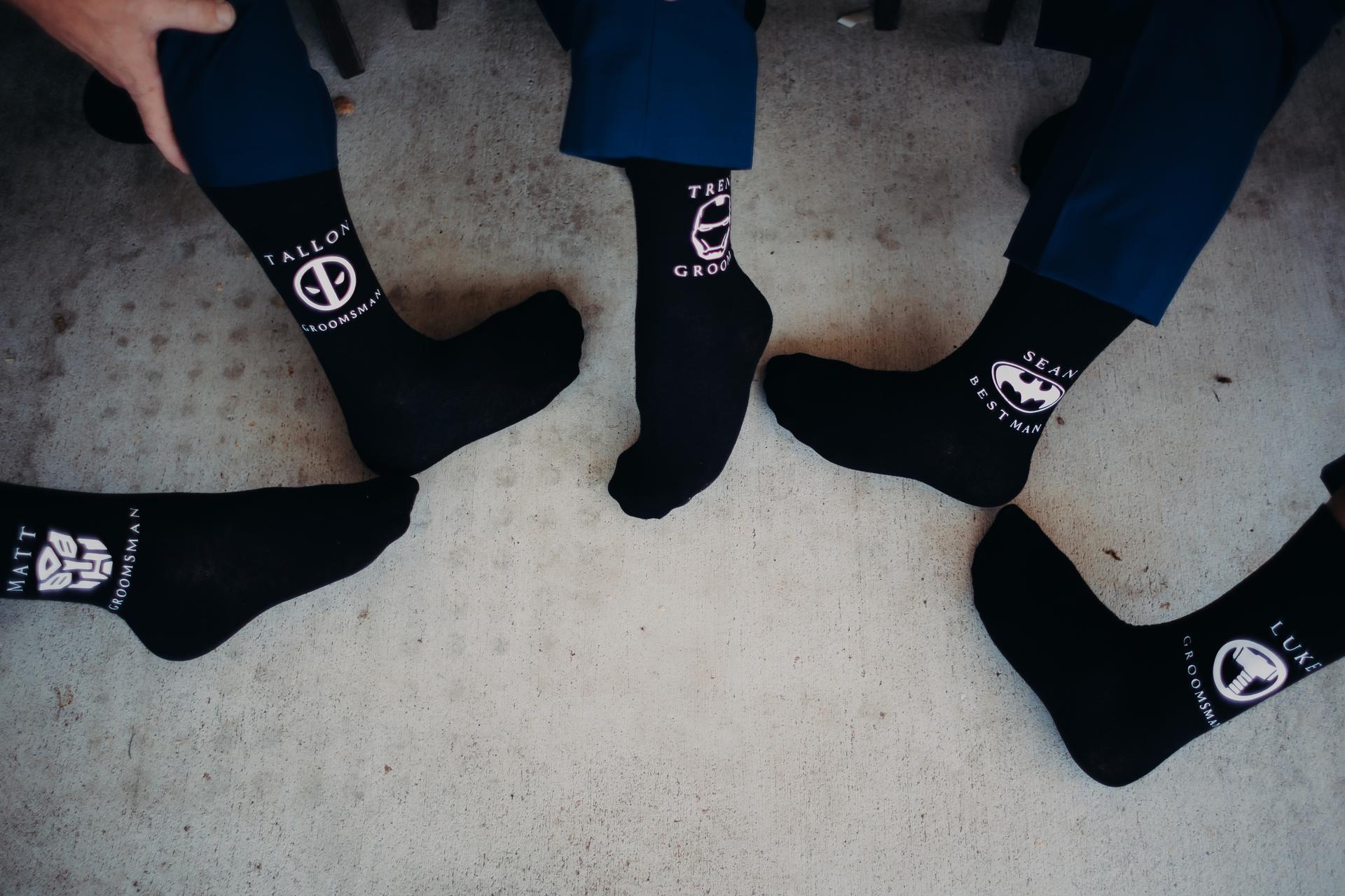 Five men wearing black socks with personalized superhero logos and names, including 'Trent Groom', 'Sean Best Man', and other groomsmen, showing their feet on a concrete floor at Ocean View Estates.