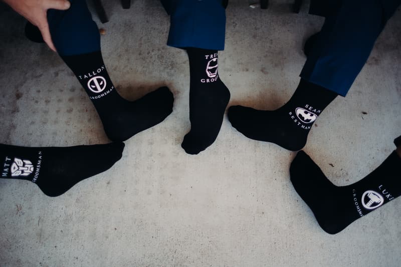 Five men wearing black socks with personalized superhero logos and names, including 'Trent Groom', 'Sean Best Man', and other groomsmen, showing their feet on a concrete floor at Ocean View Estates.