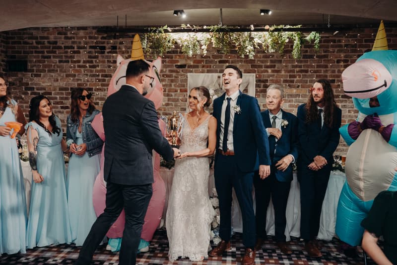 Courtney the bride and Cameron the groom stand together at the Sandstone Point Hotel — Cellar reception stage, receiving a trophy from a man in a dark suit. Bridesmaids in light blue dresses and two people in inflatable unicorn costumes stand nearby, along with two men in suits, one older and one younger.
