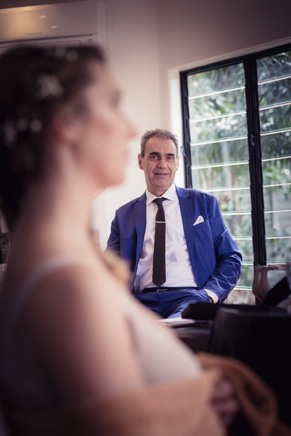 The bride is in the foreground out of focus, while an older man in a blue suit and black tie, likely the bride's father, is seated and looking toward her near a window with metal bars.