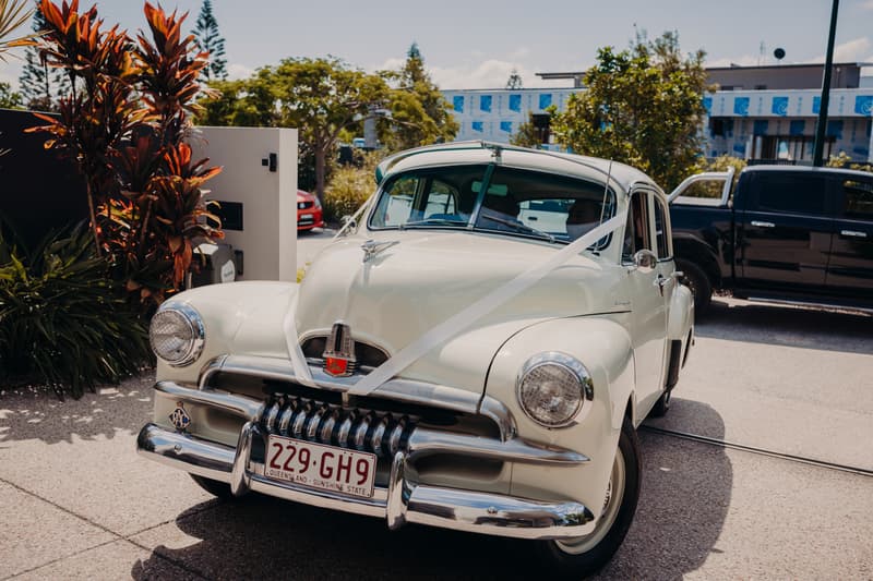 A vintage white car decorated with a white ribbon is parked outside near foliage and a building under construction.