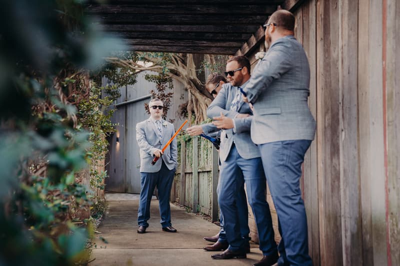 The groom and three groomsmen dressed in light grey jackets and blue pants stand under a wooden pergola at Sandstone Point Hotel Rustic Arbour, with the groom holding a toy lightsaber.