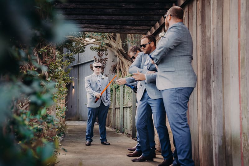 The groom and three groomsmen dressed in light grey jackets and blue pants stand under a wooden pergola, with the groom holding a toy lightsaber. Shortest day wedding timeline by She Said Yes.
