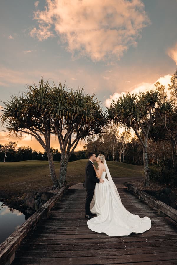 Bride Libby and groom Kyle stand on a wooden bridge at The Tides, sharing a kiss at sunset with trees and a grassy hill in the background.