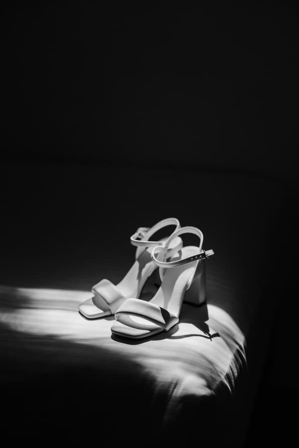 A pair of white bridal high-heeled sandals placed on a surface with dramatic lighting highlighting them.