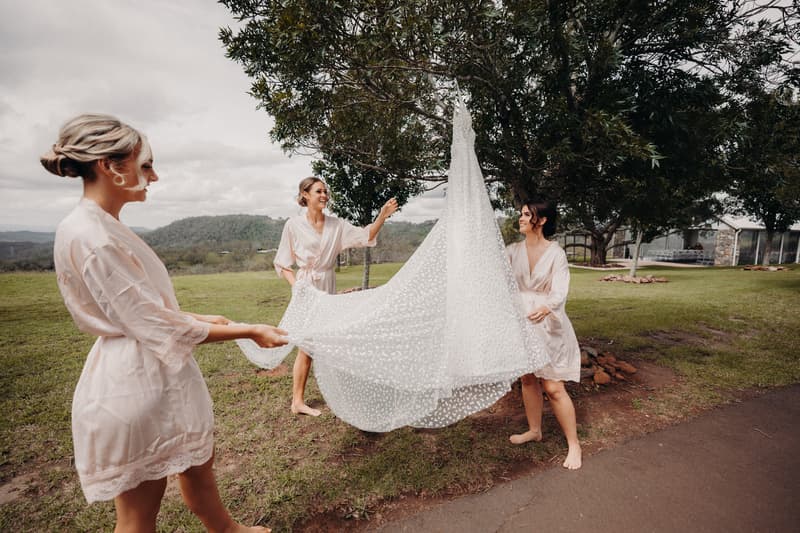 Three bridesmaids in matching robes hold up a wedding dress outdoors at Preston Peak Winery Functions Centre with trees and hills in the background.