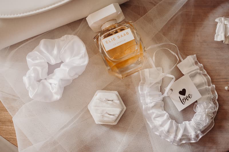 Wedding accessories including a white satin scrunchie, a bottle of Elie Saab Le Parfum, two wedding rings in a white hexagonal ring box, and a white garter with a tag that reads 'love' are arranged on a beige tulle fabric.