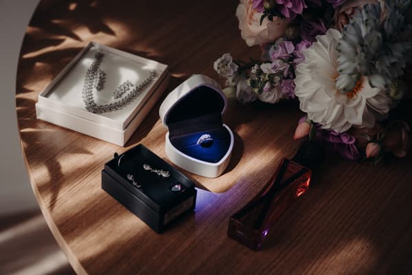 Close-up of wedding jewelry including a ring in a white box, two sets of earrings and a necklace in black and white boxes, a perfume bottle, and a bridal bouquet on a wooden table at Sandstone Point Hotel.