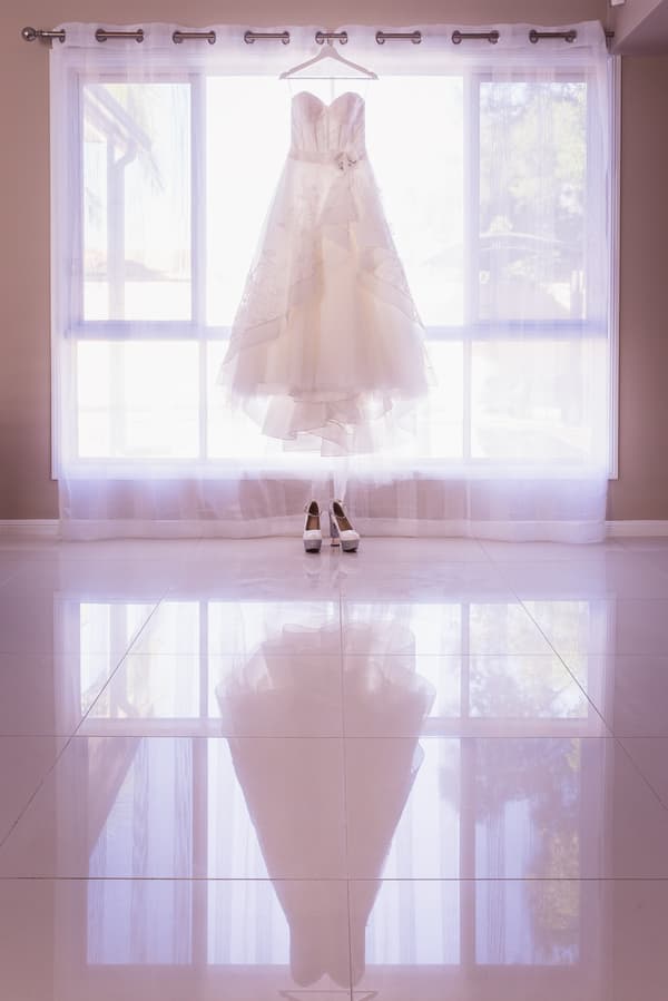 The bride's wedding dress is hanging on a curtain rod in front of a large window at Kwila Lodge, with a pair of white bridal shoes placed on the floor beneath it.