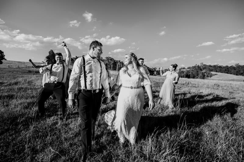 Bride Annie and groom Trent walk hand in hand through a grassy field at Ocean View Estates, followed by bridesmaids and groomsmen in celebratory poses.