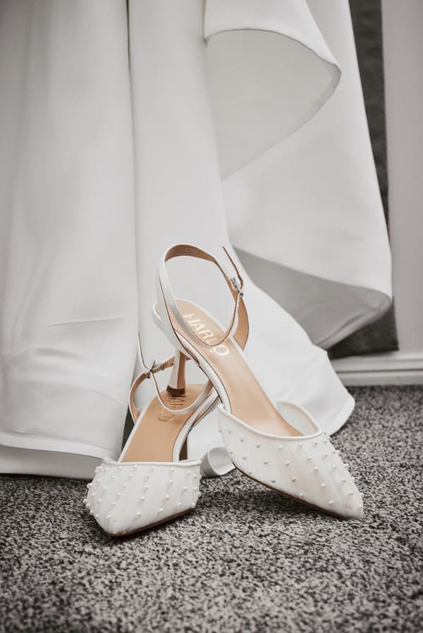 Close-up of the bride's white wedding shoes with pearl embellishments placed on carpeted floor next to the hem of the wedding dress.