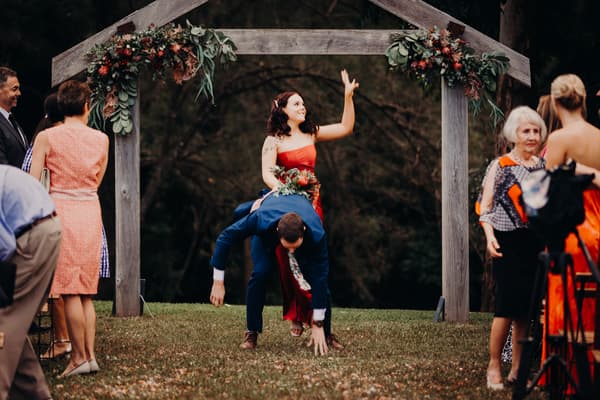The bride and groom are under a wooden floral arch at Yabbaloumba Retreat — By The River, with the groom bent over and the bride sitting on his back waving. Wedding guests stand nearby watching.
