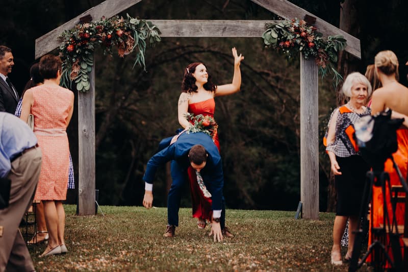 The bride and groom are under a wooden floral arch at Yabbaloumba Retreat — By The River, with the groom bent over and the bride sitting on his back waving. Wedding guests stand nearby watching.