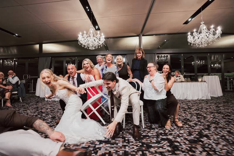 The bride Morgan and groom Joel play a group game with guests at the reception in The Pumicestone Room, Sandstone Point Hotel, with guests seated and laughing behind them.