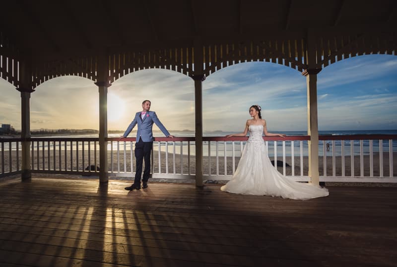 The bride Wing in a strapless white wedding gown and the groom Jason in a blue suit with a pink bow tie pose separately under a wooden pavilion overlooking the beach at Bilinga Beach Weddings during sunset.