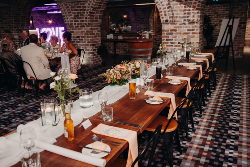 Reception tables set with glassware, plates, cutlery, floral centerpieces, and drinks at Sandstone Point Hotel — Cellar, with guests seated and a neon LOVE sign visible in the background.