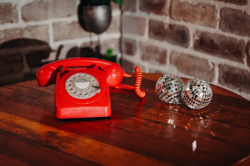 A red rotary dial telephone and two small disco balls are placed on a wooden table against a brick wall at Sandstone Point Hotel — Cellar.