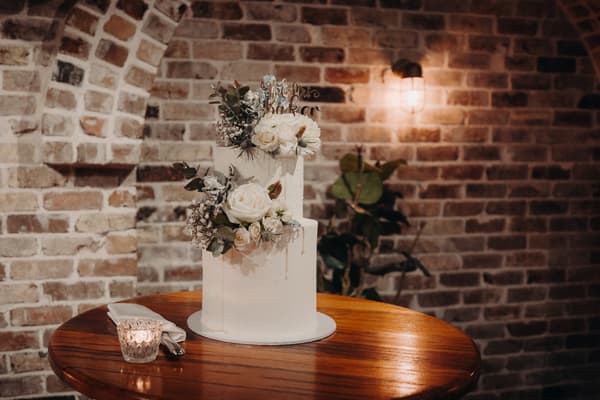 Two-tier white wedding cake decorated with white flowers and greenery on a wooden table at Sandstone Point Hotel — Cellar, with a lit candle and napkin with cutlery beside it.