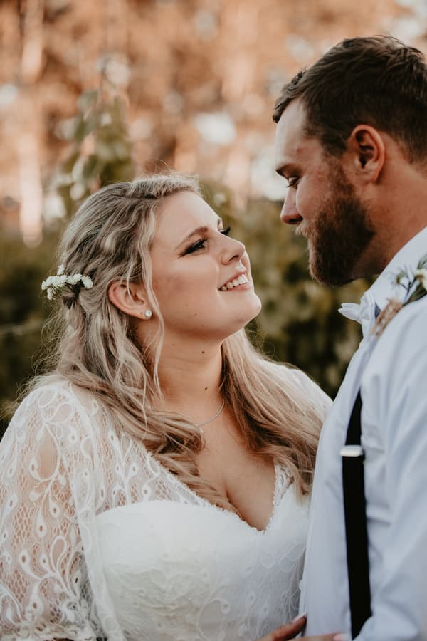 Annie the bride and Trent the groom face each other closely outdoors at Ocean View Estates, with Annie smiling and wearing a lace wedding dress and floral hair accessory, and Trent in a white shirt with suspenders and boutonniere.