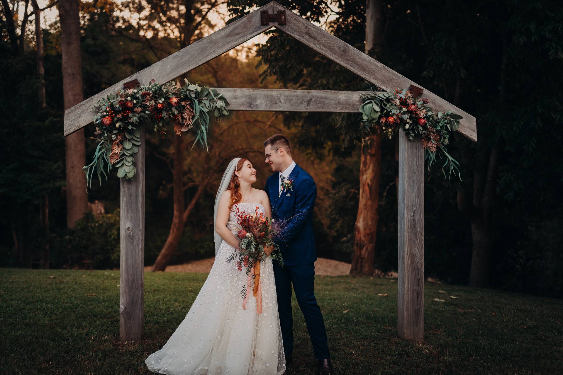 Lilly and Connor stand together under a wooden floral arch at Yabbaloumba Retreat, Yabbaloumba Retreat, posing for couple portraits with trees and grass in the background.