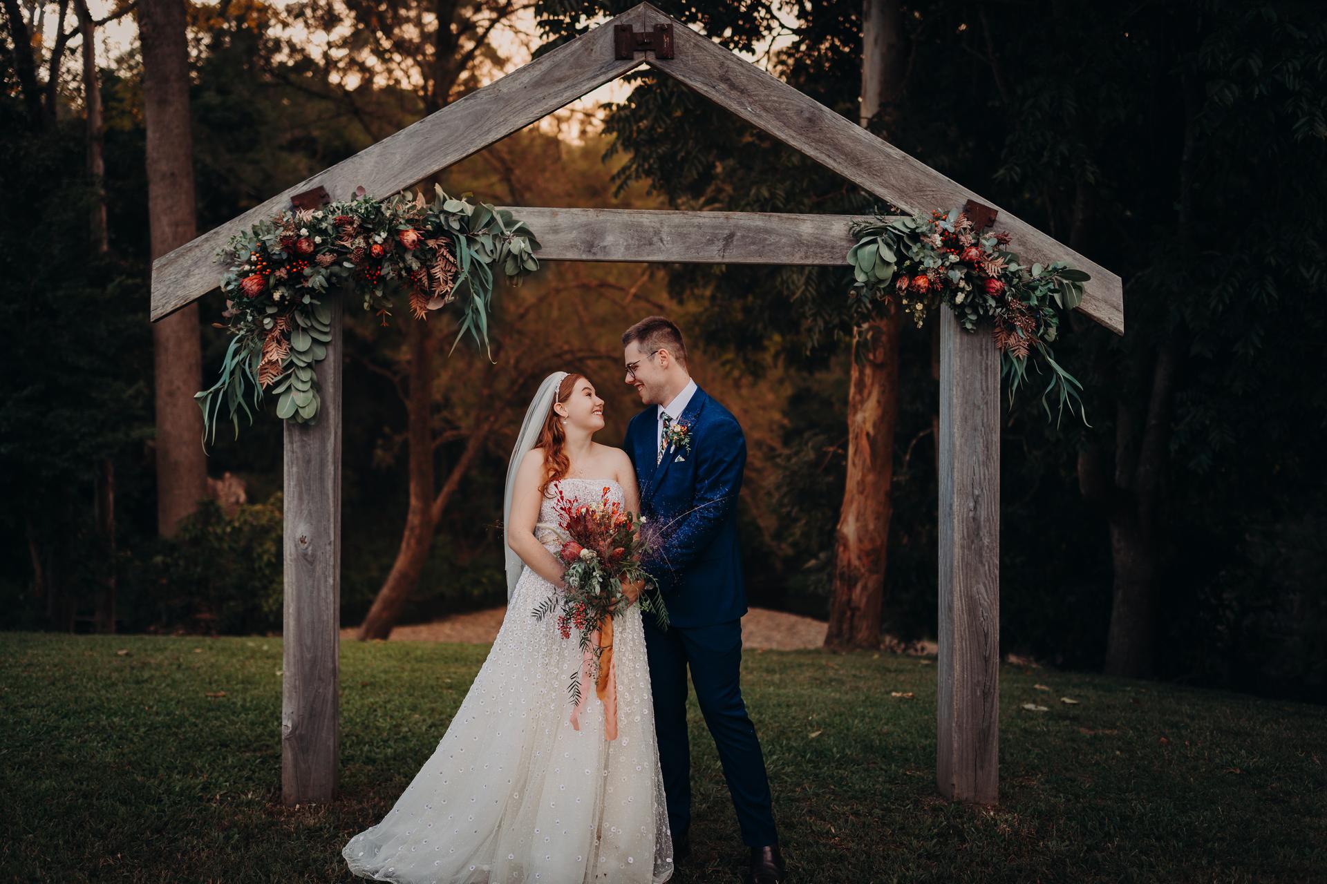 Lilly and Connor stand together under a wooden floral arch at Yabbaloumba Retreat, Yabbaloumba Retreat, posing for couple portraits with trees and grass in the background.