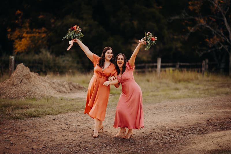 Two bridesmaids in orange and pink dresses pose outdoors holding bouquets at Yabbaloumba Retreat.