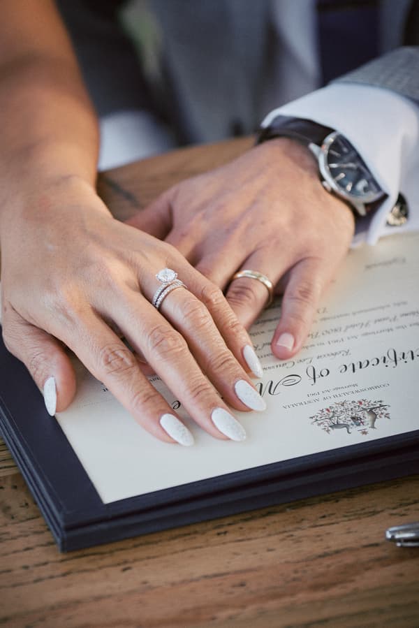 The bride and groom's hands rest on a marriage certificate at Sandstone Point Hotel — The Pavilion, showing the bride's engagement and wedding rings and the groom's wedding band.