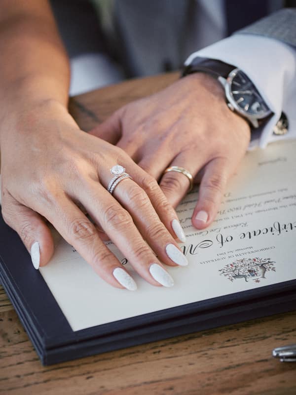 The bride and groom's hands rest on a marriage certificate at Sandstone Point Hotel — The Pavilion, showing the bride's engagement and wedding rings and the groom's wedding band.