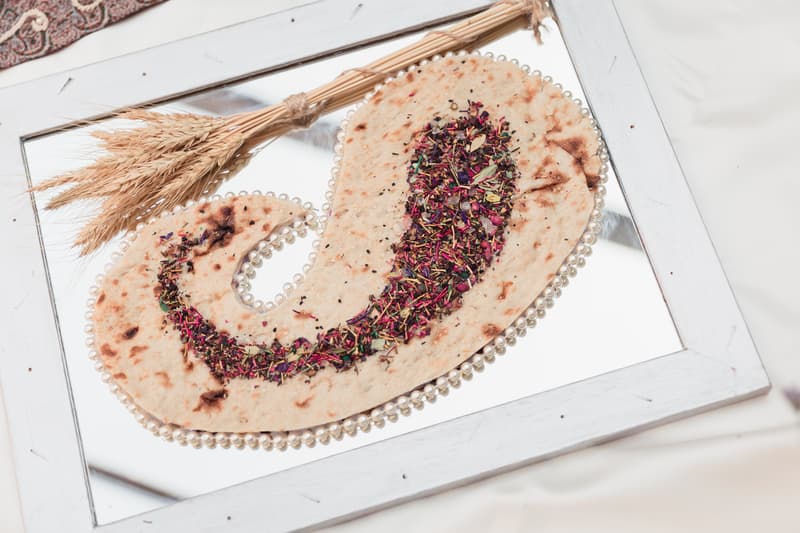 A decorative flatbread with colorful dried herbs arranged in a paisley pattern on a mirror tray with a white wooden frame, accompanied by a small bundle of dried wheat stalks.